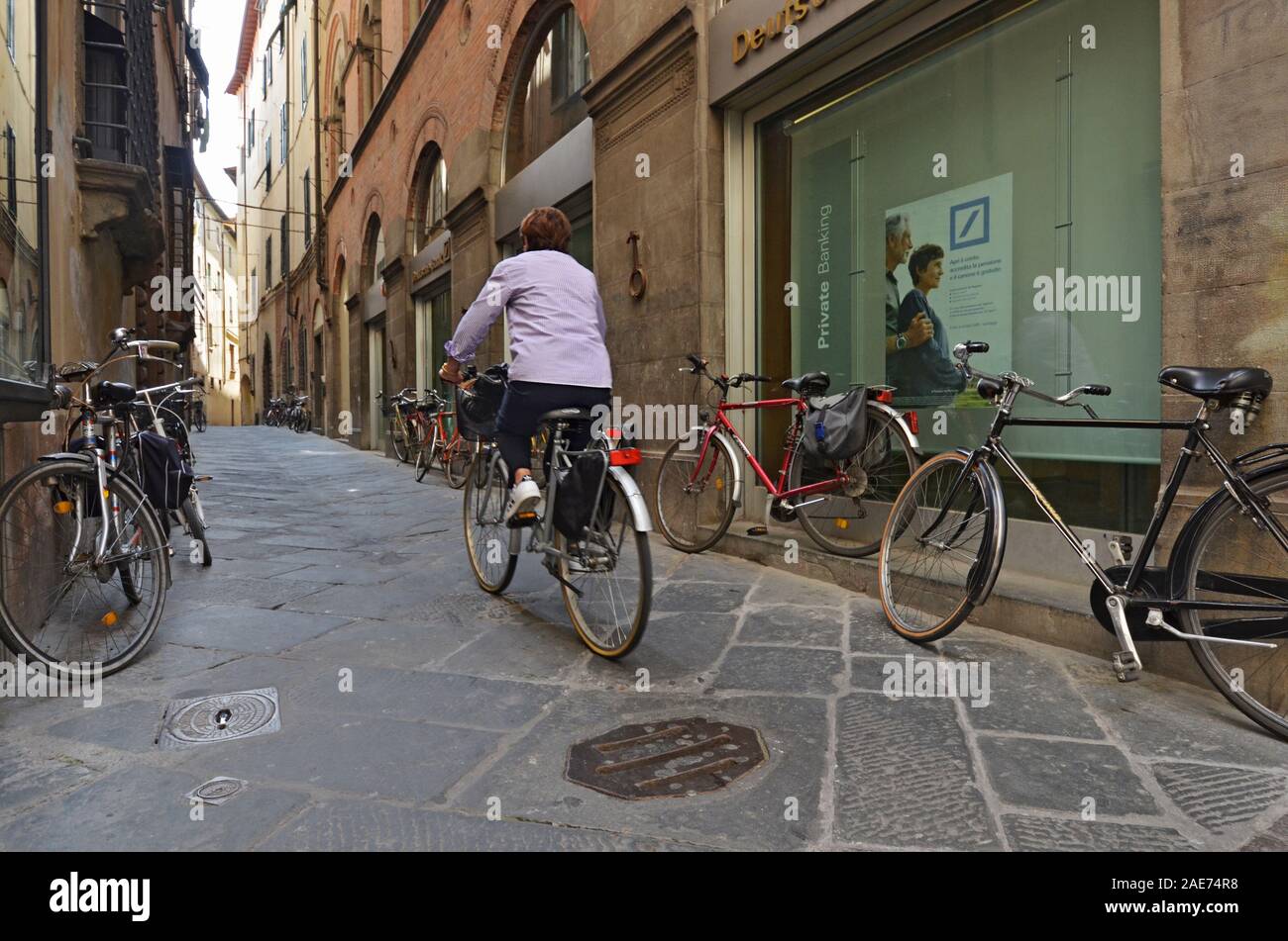 A middle aged woman rides her bicycle down a cobblestone street in the Italian city of Lucca. Stock Photo