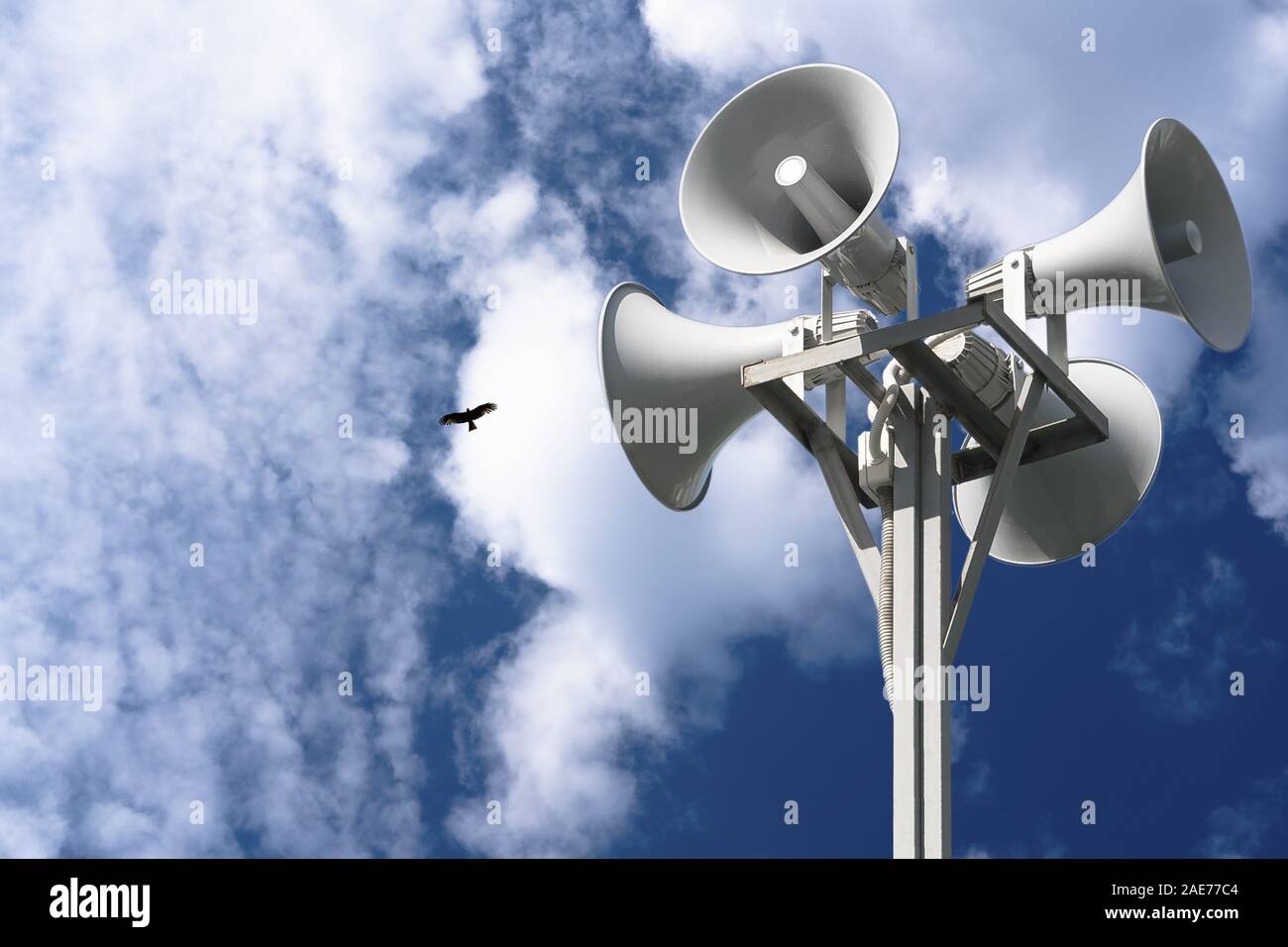 A column with four gray loudspeakers in a circle against a background of clouds and a flying bird. Hazard warning system. Ability to post your test or Stock Photo