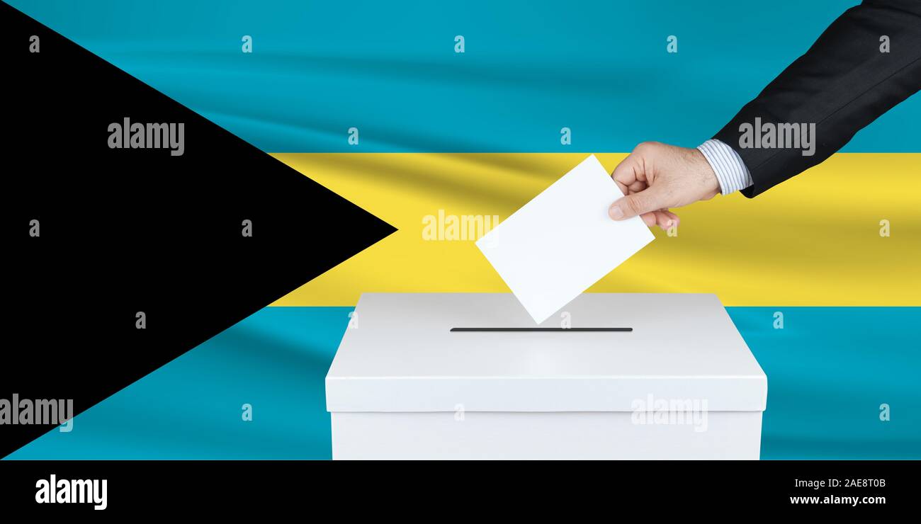 Election in Bahamas. The hand of man putting his vote in the ballot box. Waved Bahamas flag on background. Stock Photo