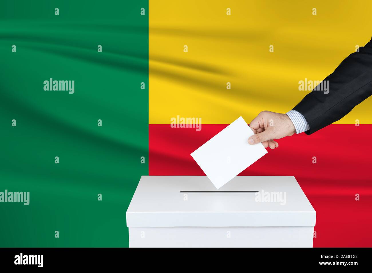Election in Benin. The hand of man putting his vote in the ballot box. Waved Benin flag on background. Stock Photo