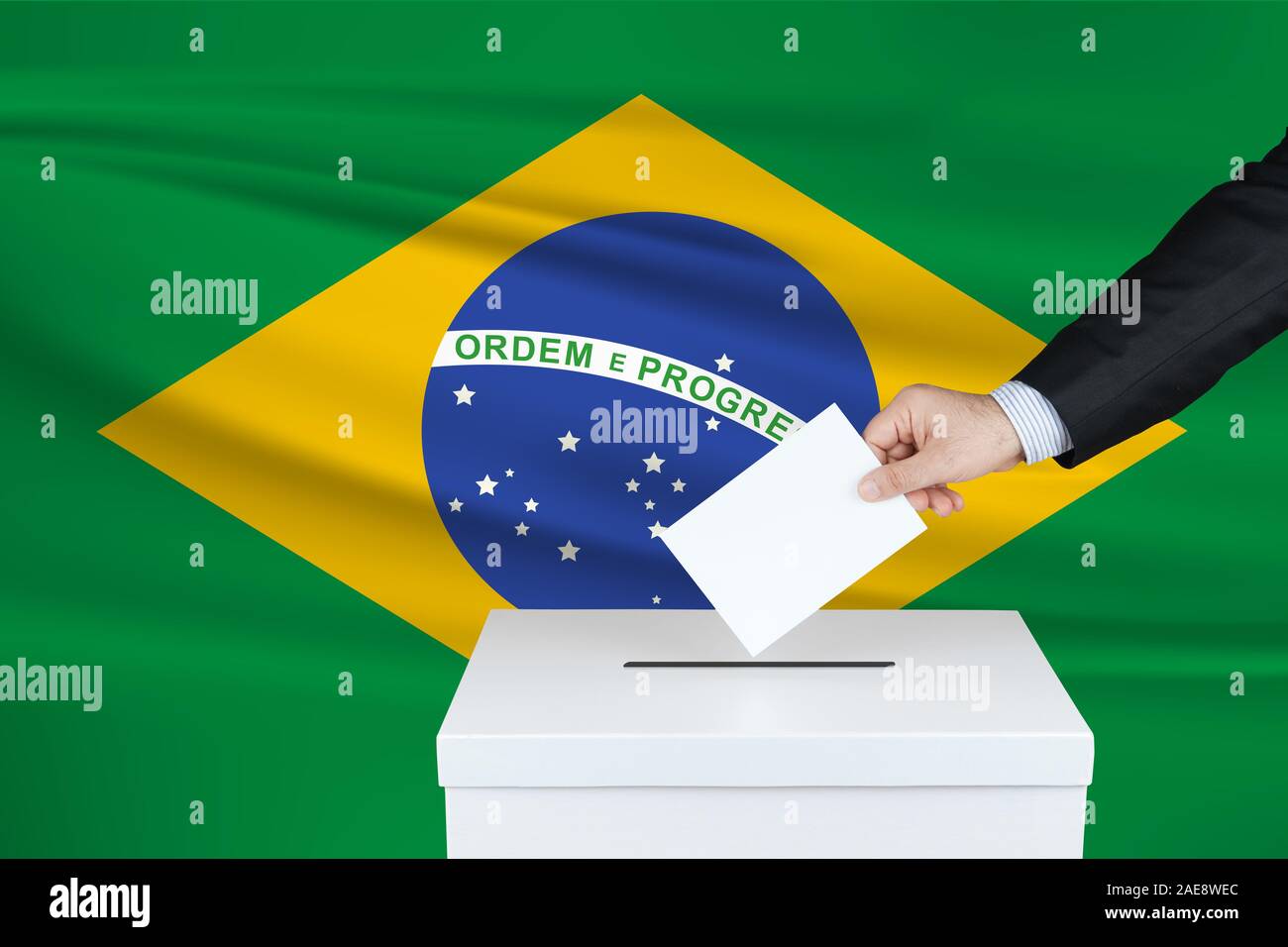 Election in Brazil. The hand of man putting his vote in the ballot box. Waved Brazil flag on background. Stock Photo