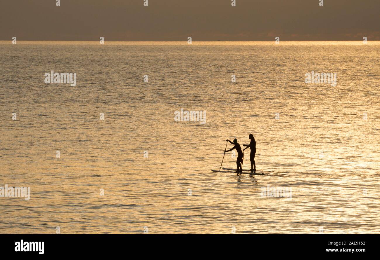 A woman and a man using stand up paddleboards outside Lyme Regis harbour in early December at sunrise. Lyme Regis is situated on the heritage coast or Stock Photo
