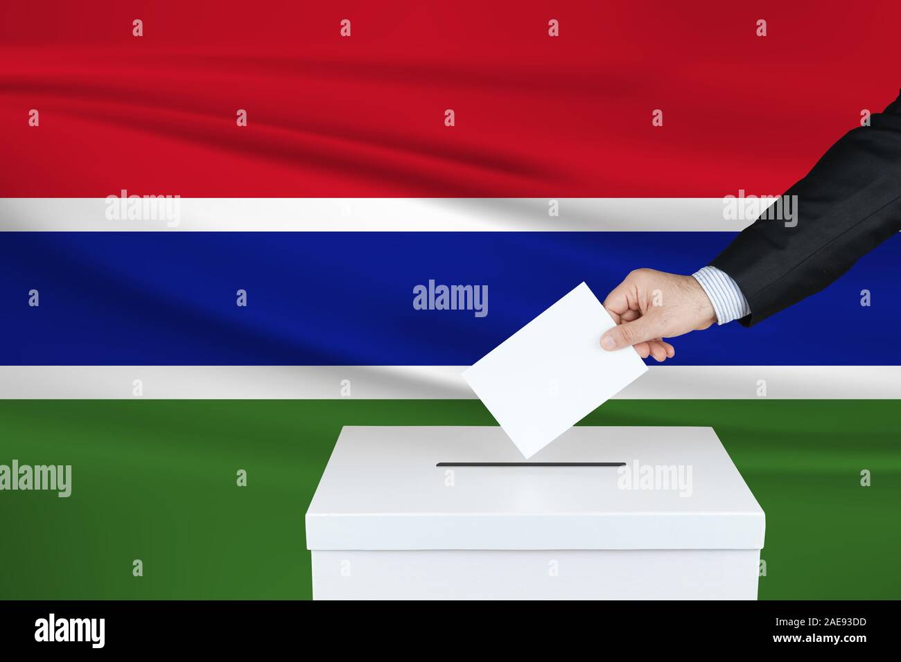 Election in Gambia. The hand of man putting his vote in the ballot box. Waved Gambia flag on background. Stock Photo