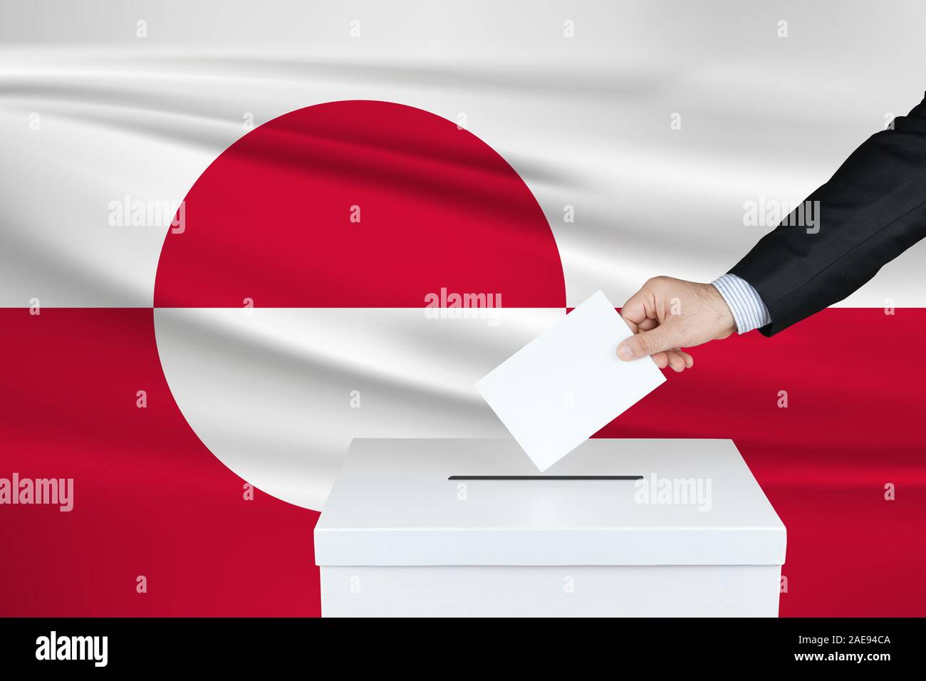 Election in Greenland. The hand of man putting his vote in the ballot box. Waved Greenland flag on background. Stock Photo