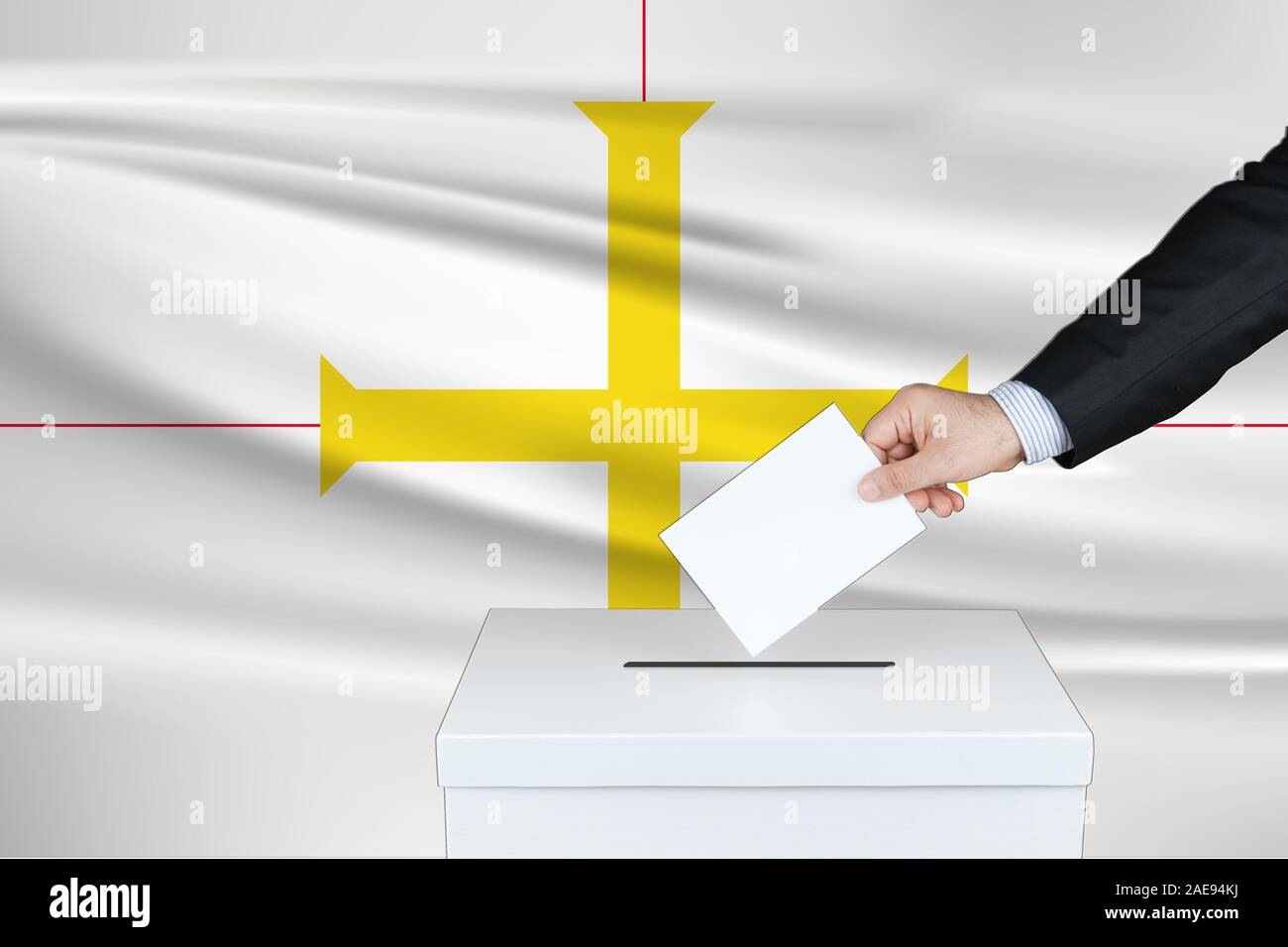 Election in Guernsey. The hand of man putting his vote in the ballot box. Waved Guernsey flag on background. Stock Photo