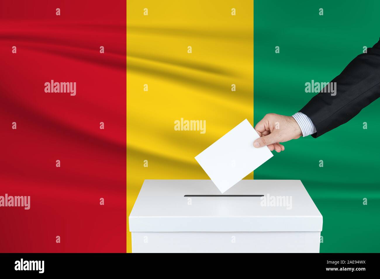 Election in Guinea. The hand of man putting his vote in the ballot box. Waved Guinea flag on background. Stock Photo