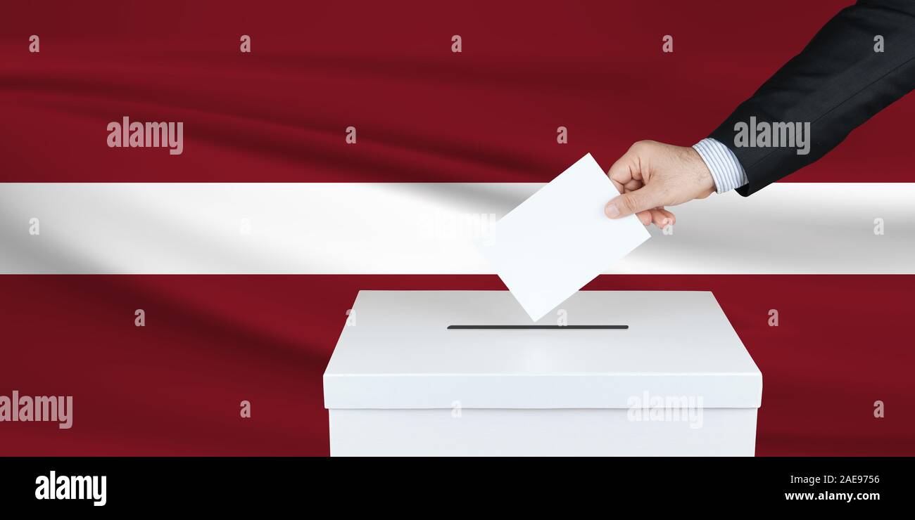Election in Latvia. The hand of man putting his vote in the ballot box. Waved Latvia flag on background. Stock Photo