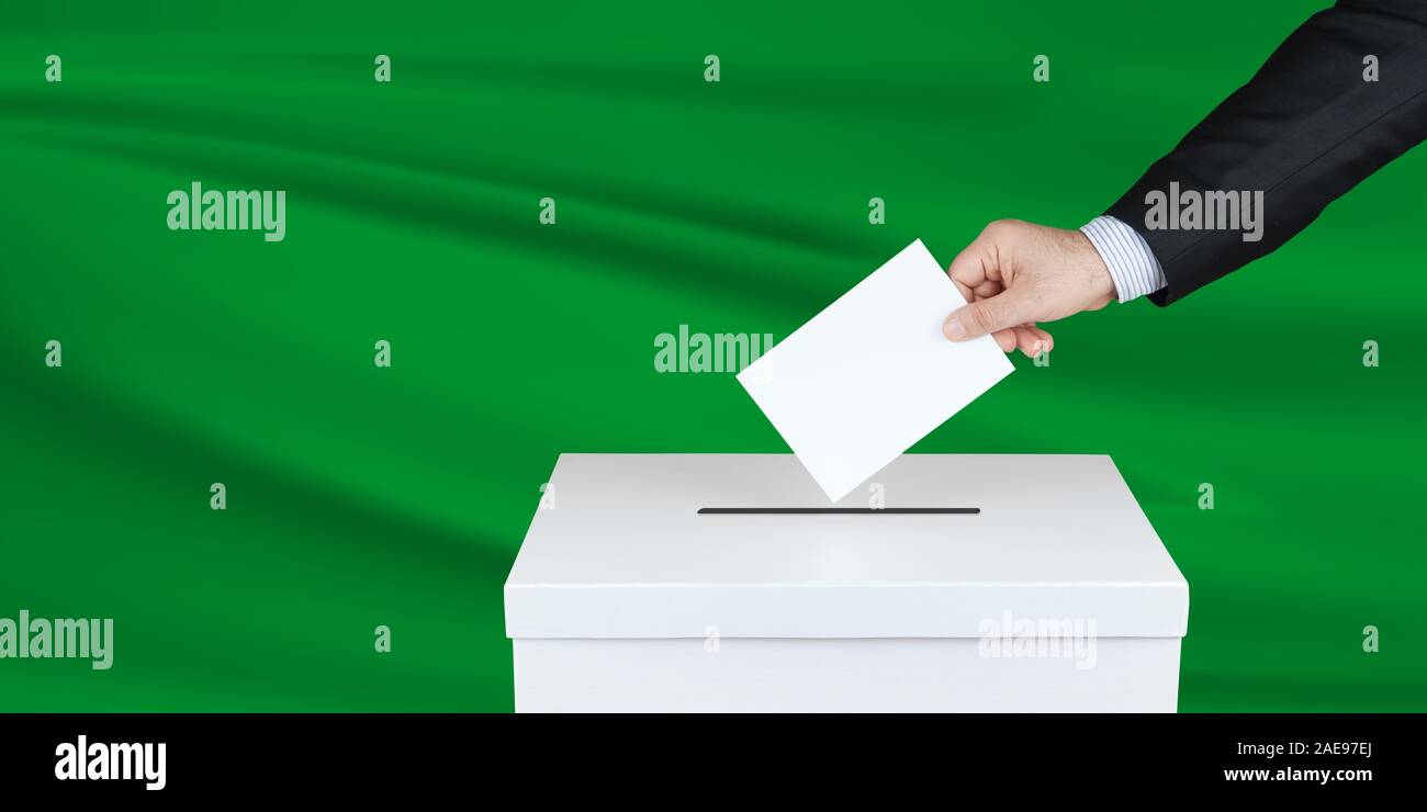 Election in Libya. The hand of man putting his vote in the ballot box. Waved Libya flag on background. Stock Photo