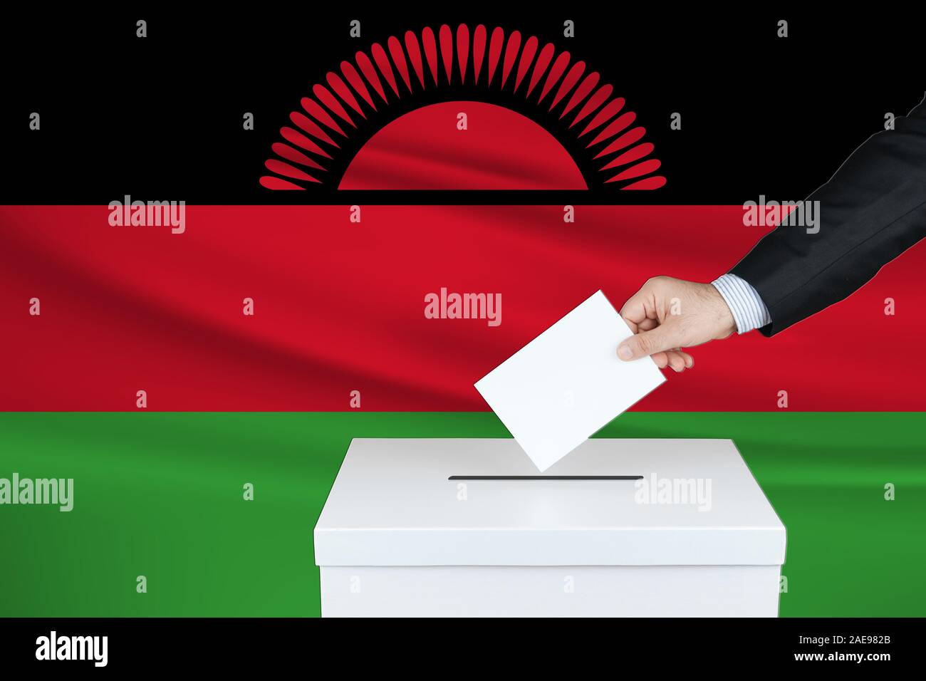 Election in Malawi. The hand of man putting his vote in the ballot box. Waved Malawi flag on background. Stock Photo