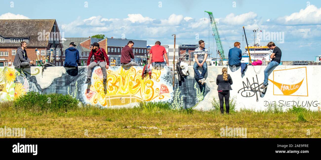 Woman talking to a group of young men sitting on a wall covered in graffiti drinking beers in the port of Cuxhaven Lower Saxony Germany. Stock Photo