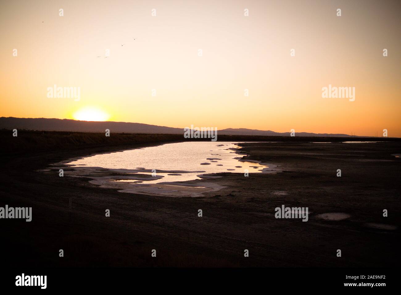 Sun setting behind mountain, with tidal marsh / salt pan on the edge of San Fransisco Bay in the foreground. Orange sky, black ground. Stock Photo