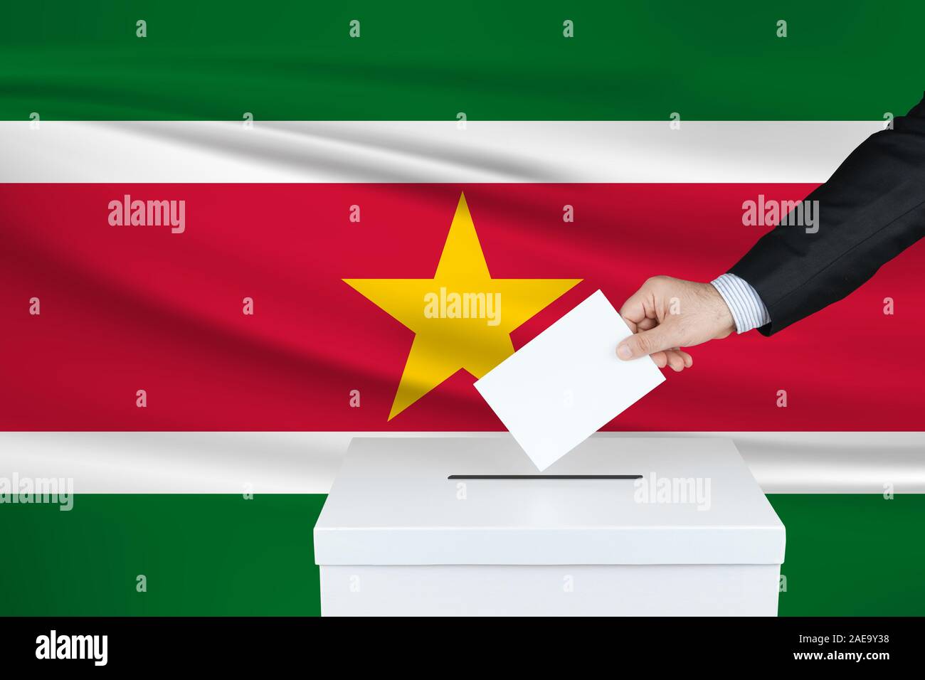 Election in Suriname. The hand of man putting his vote in the ballot box. Waved Suriname flag on background. Stock Photo