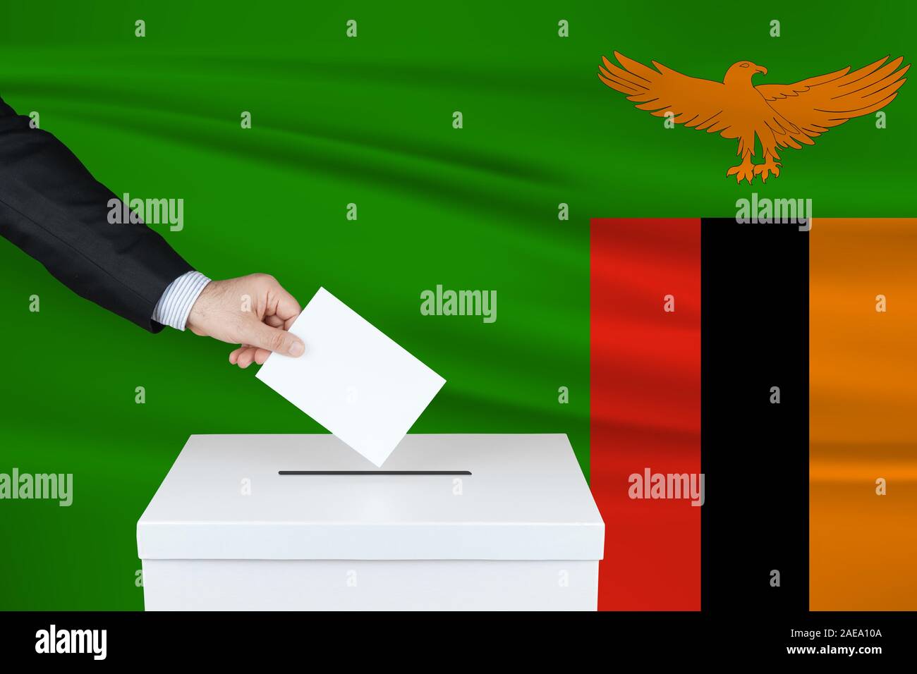 Election in Zambia. The hand of man putting his vote in the ballot box. Waved Zambia flag on background. Stock Photo