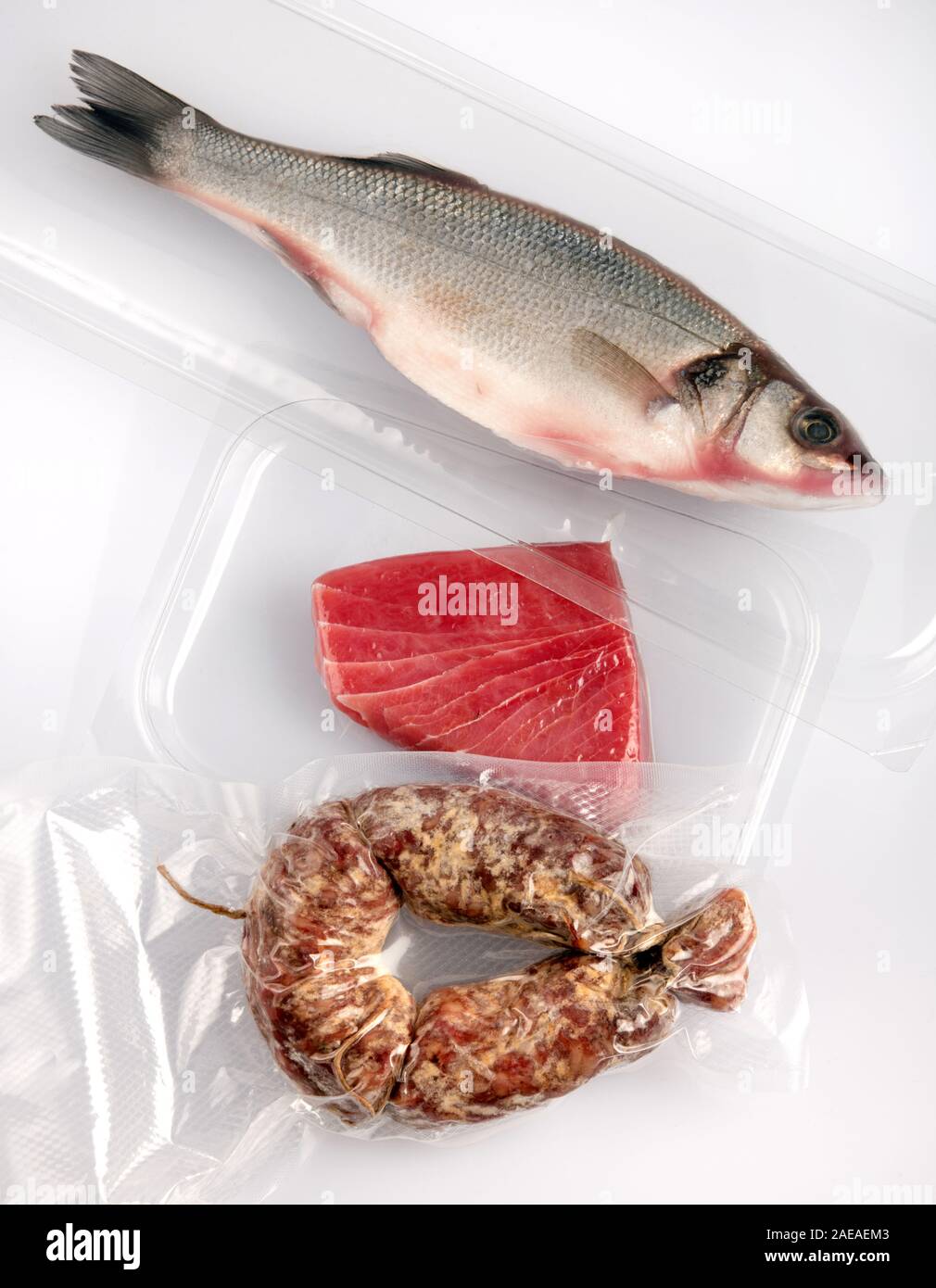 Assorted fresh foods vacuum packed in plastic for storage with a whole cleaned fish, tuna steak and spicy sausage laid out on a white table viewed fro Stock Photo