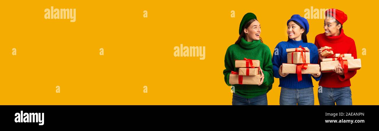 Three Girls Holding Gifts Standing In Studio Over Yellow Background Stock Photo