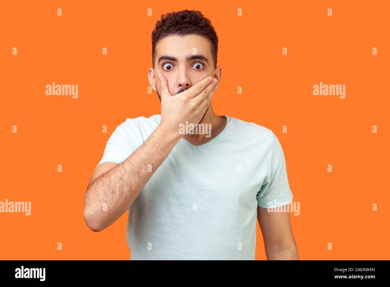 I will not say. Portrait of scared bullied man in white t-shirt covering mouth with hand, looking with frightened shocked eyes, keeping secret taboo. Stock Photo
