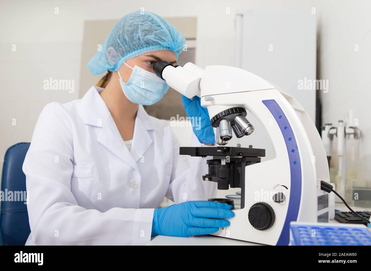 Young laboratory assistant in mask looking through modern microscope Stock Photo