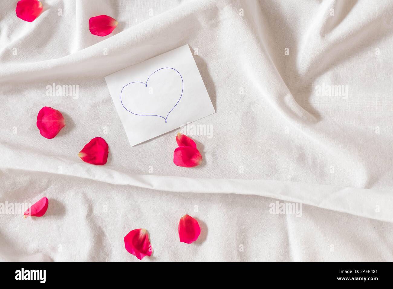 Love note with hand drawn heart on white bed sheets with pink rose petals Stock Photo