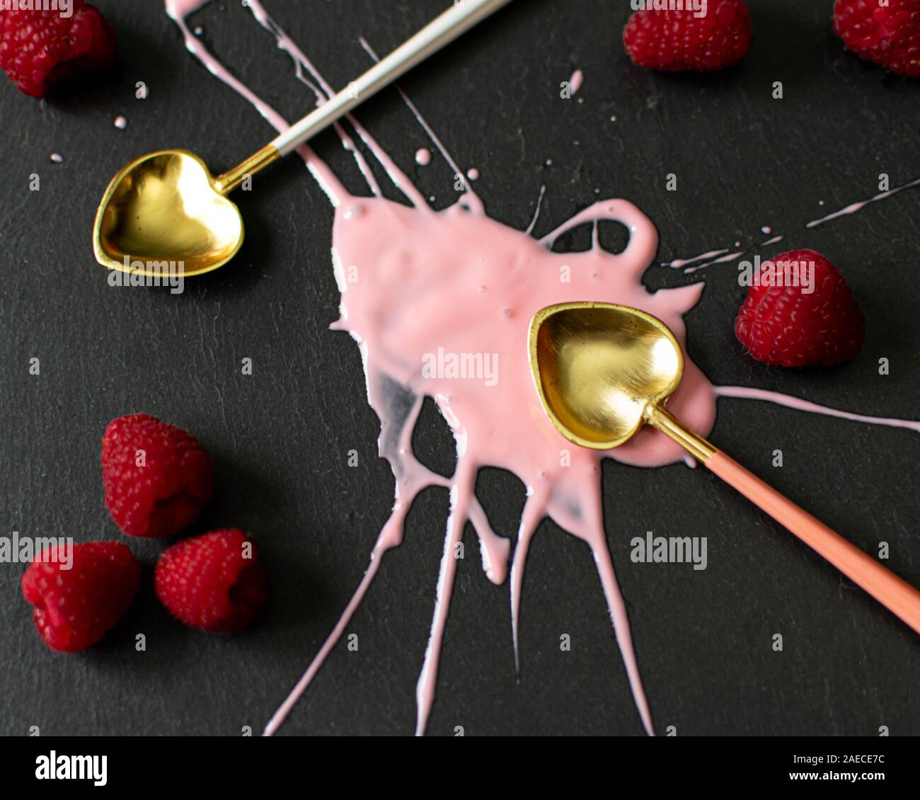 Yogurt splashed over a dark grey black background with bright pink and red raspberries and gold spoons Stock Photo
