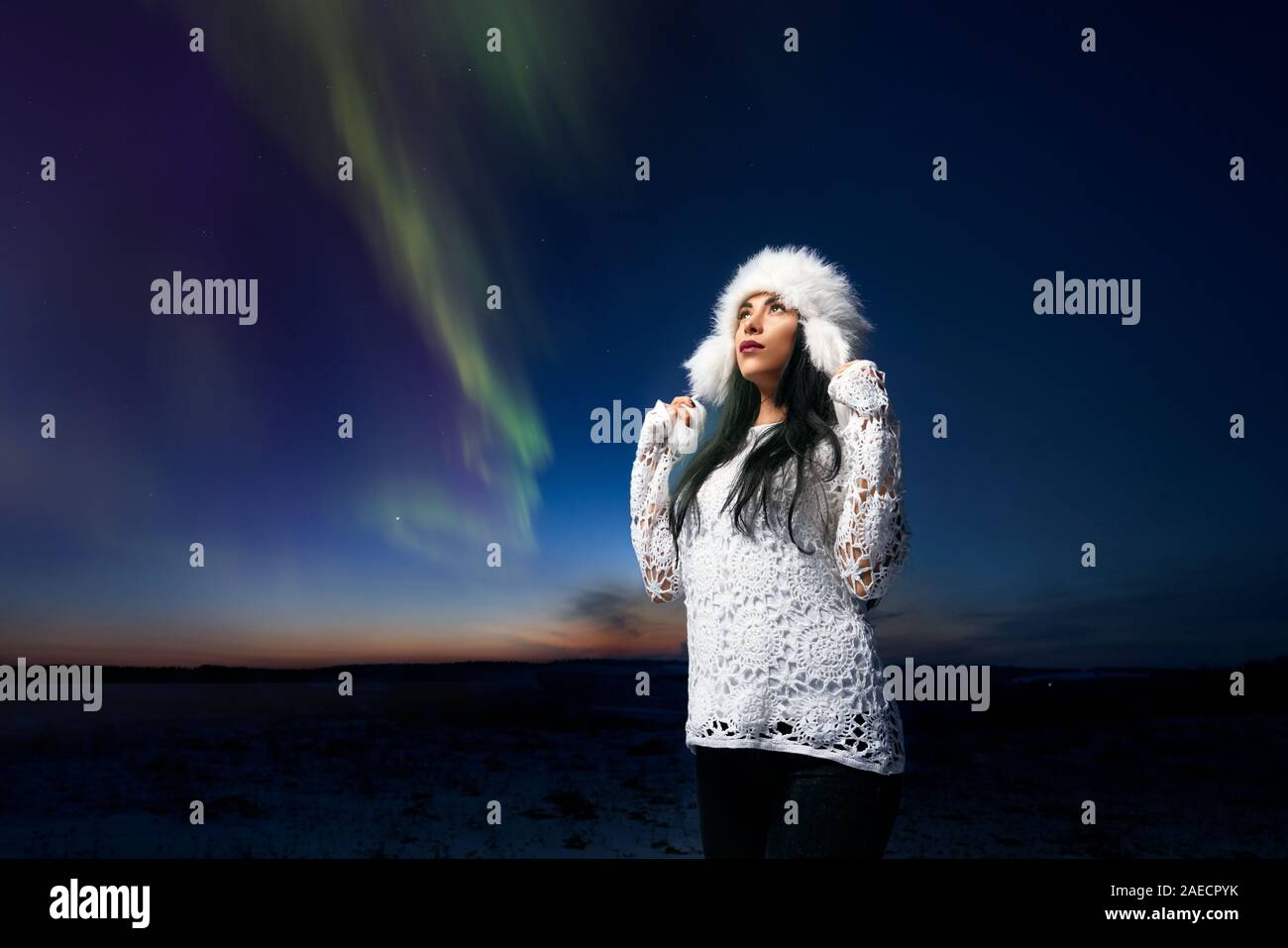 Gorgeous girl in stylish winter hat and white knitted blouse looking at night sky and admiring winter radiance. Romantic lady on background of field and bright stripes on sky. Stock Photo