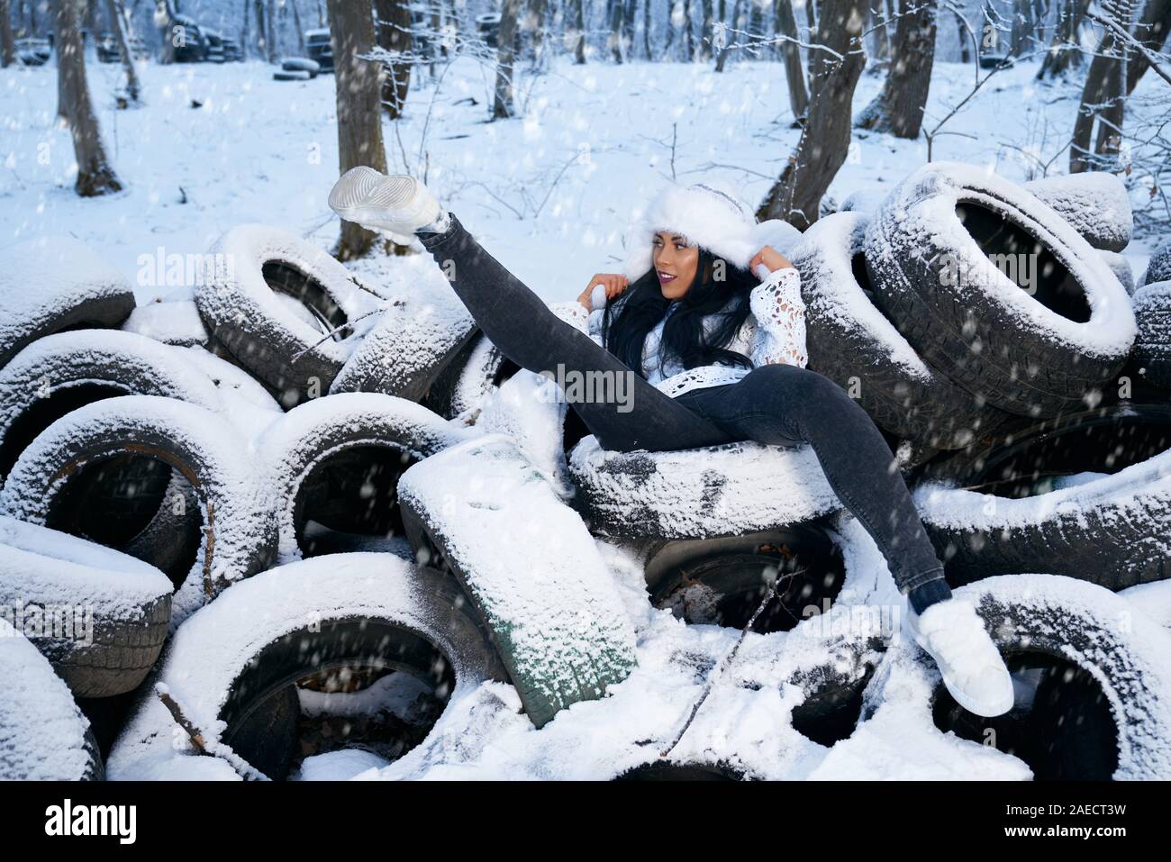 Handsome girl wearing in white warm hat, jeans and white sneakers posing on old used tires. Model lying on car tires covering with snow in forest. Lady just kidding, rejoice. Stock Photo