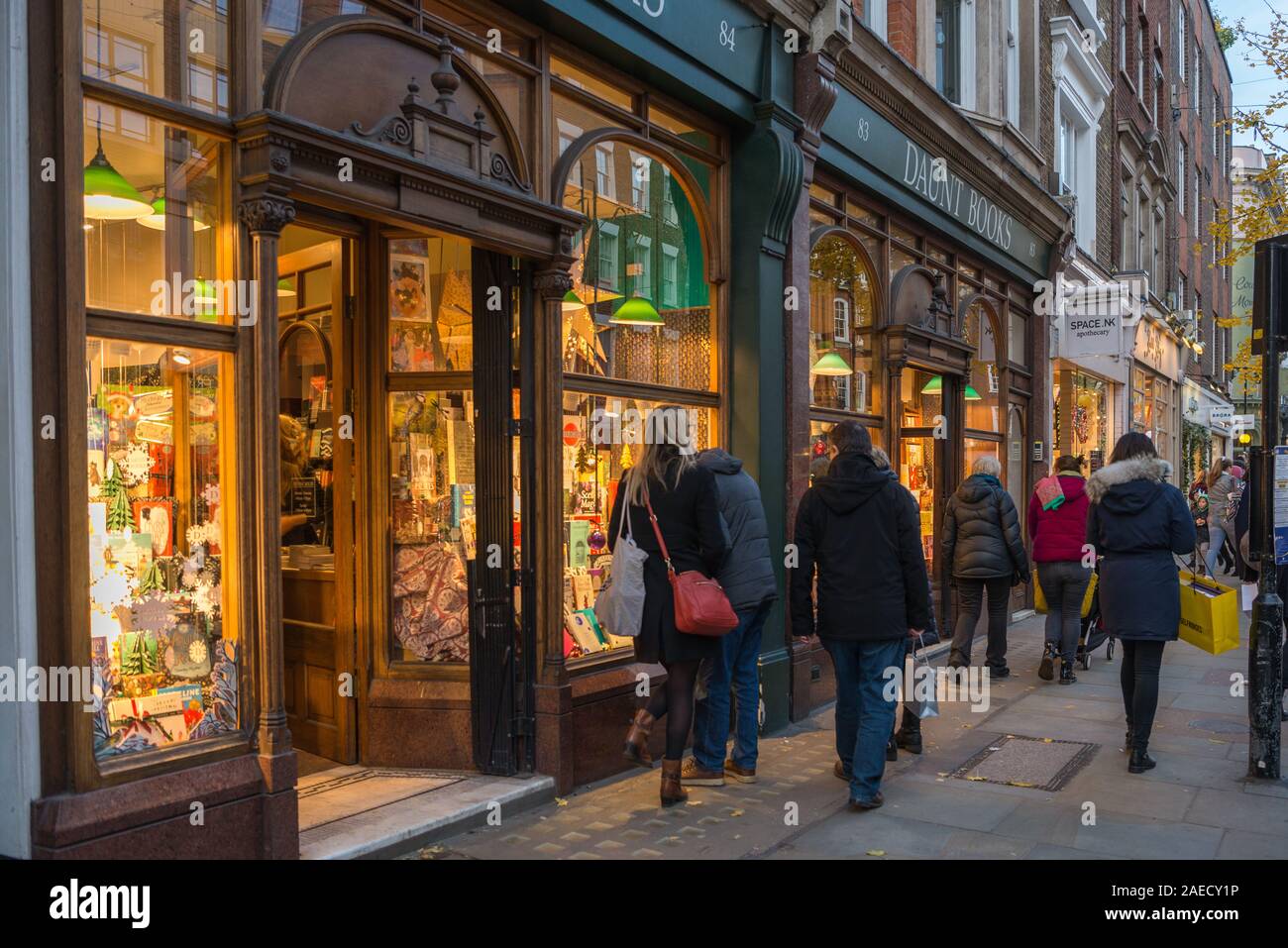 Shoppers out and about in Marylebone High Street check out the brightly lit window display at the Daunt Book Shop, London, England, UK Stock Photo