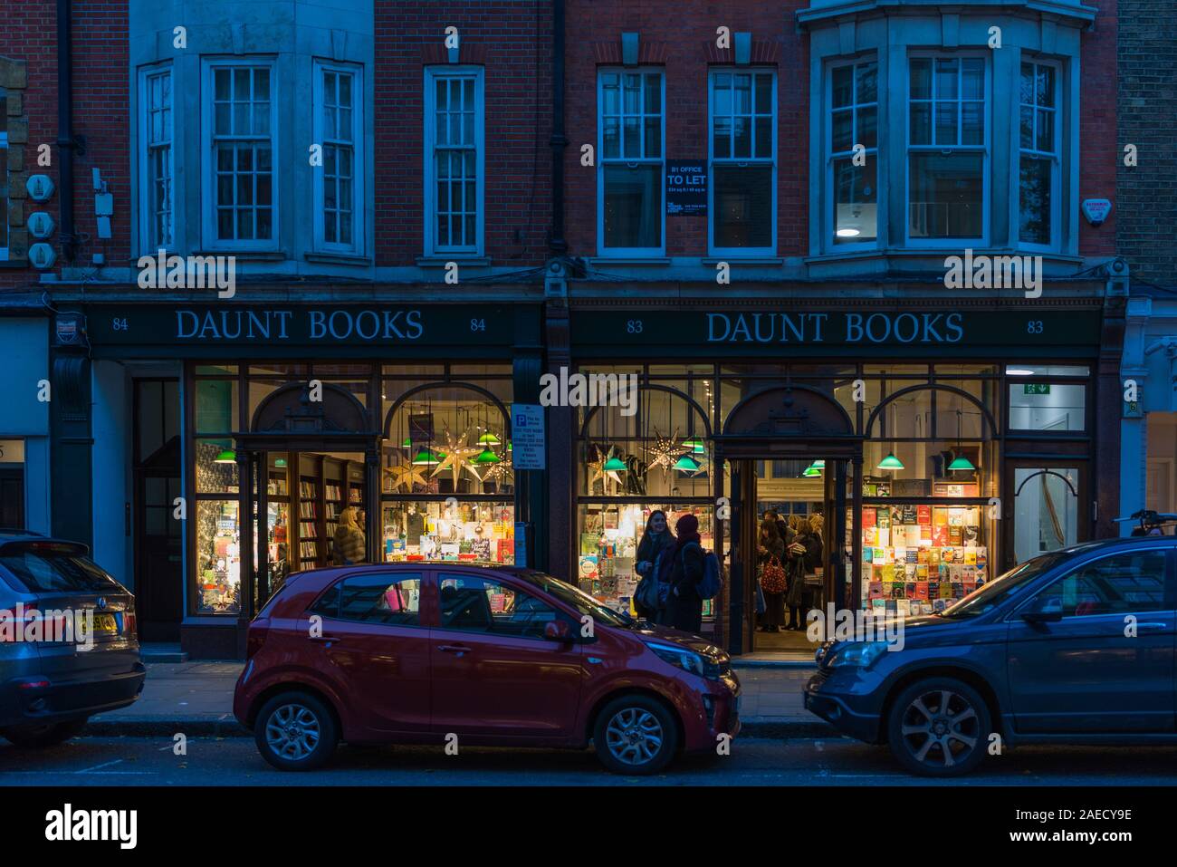 Shoppers out and about in Marylebone High Street check out the brightly lit window display at the Daunt Book Shop, London, England, UK Stock Photo