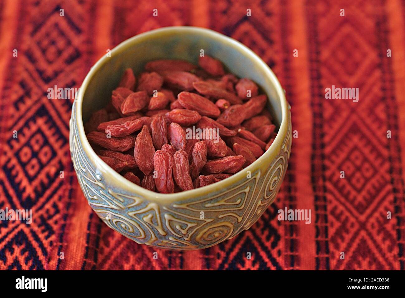 Dried Goji Berries (Lycium barbarum) in a small green ceramic bowl on a bright red tablecloth. Stock Photo