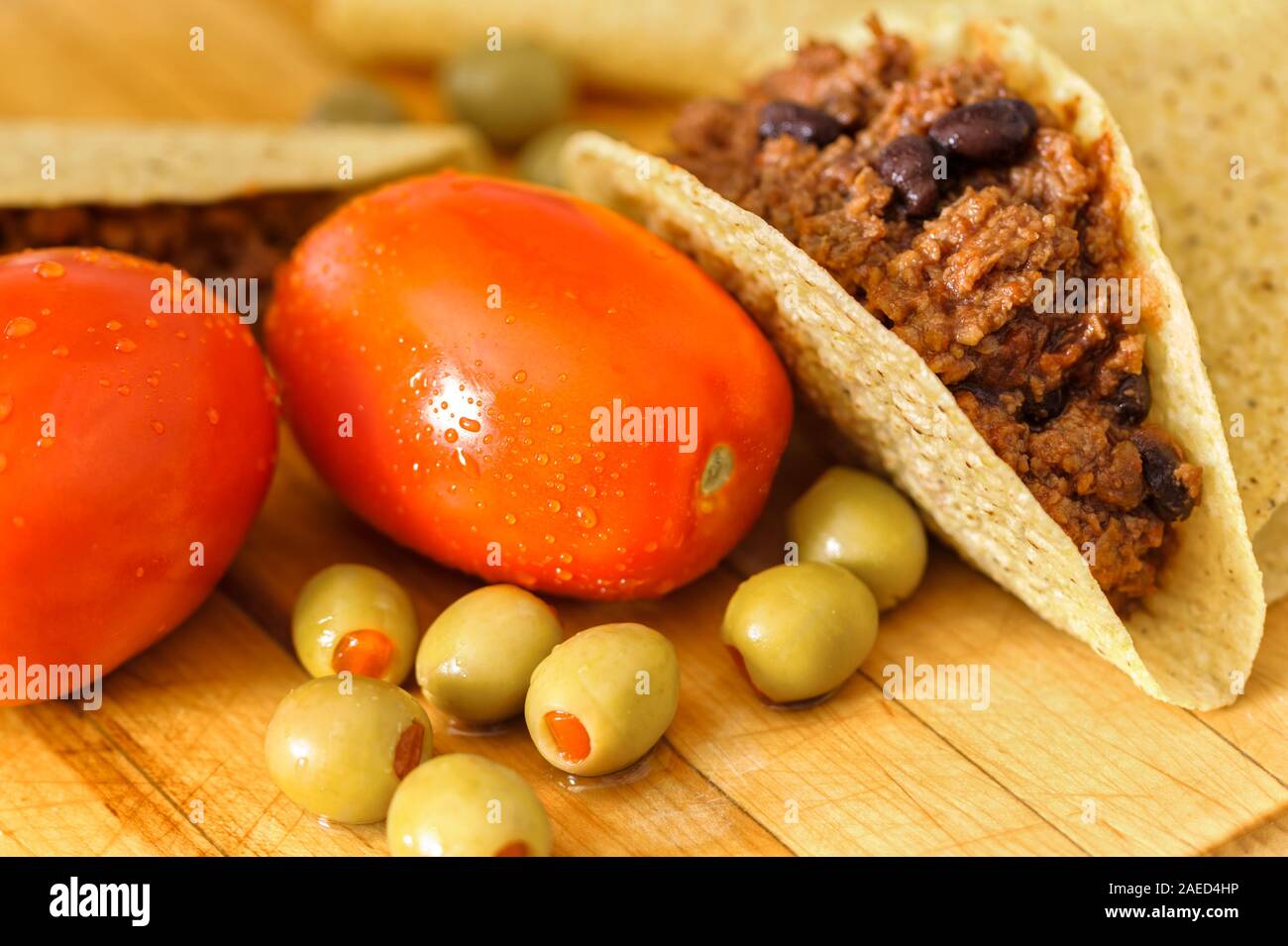 Fresh taco ingredients red ripe plum tomatoes, green olives and beef tacos with black beans in taco shells on a wooden cutting board. Stock Photo