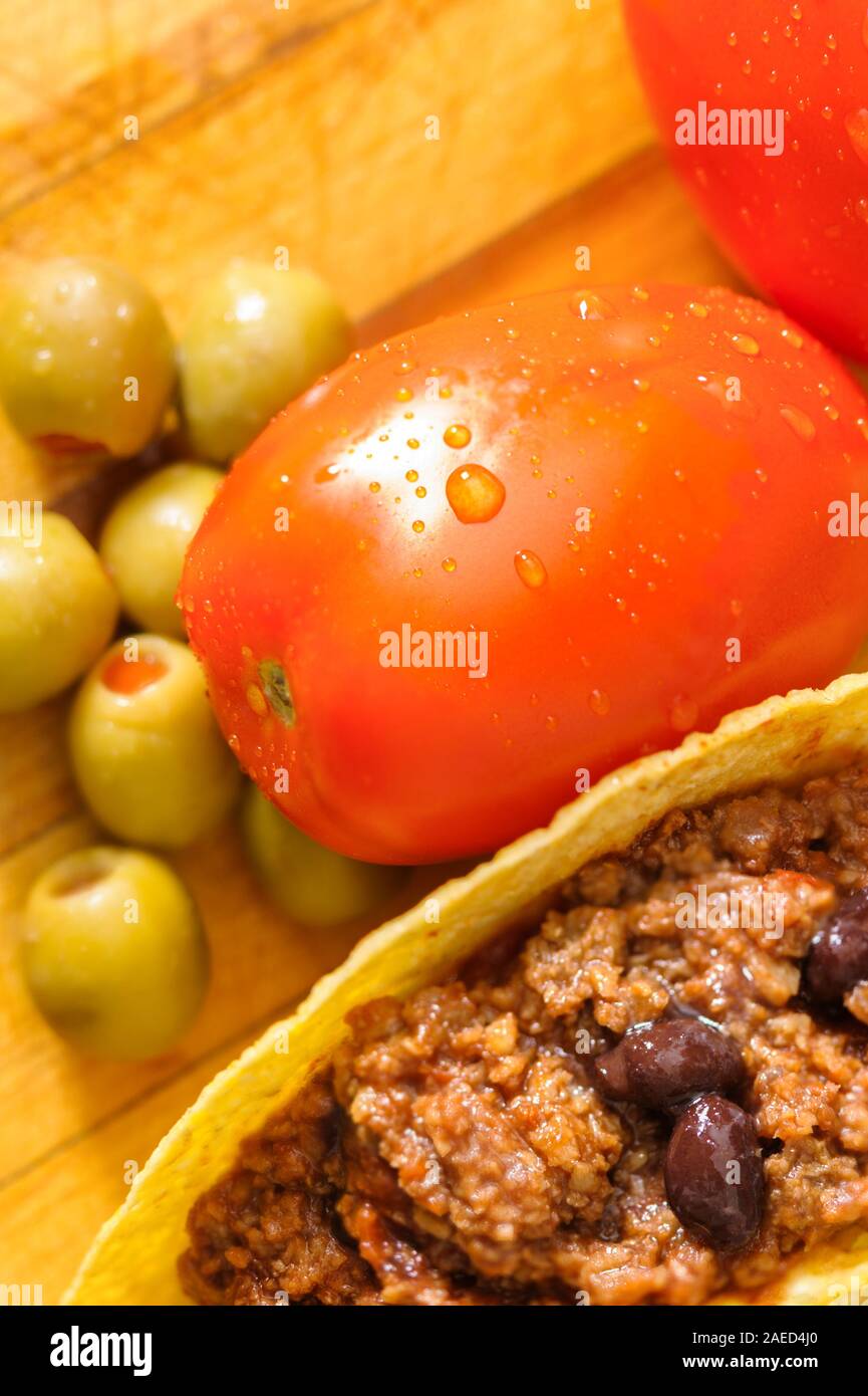 Fresh taco ingredients red ripe plum tomatoes, green olives and beef tacos with black beans in taco shells on a wooden cutting board. Stock Photo
