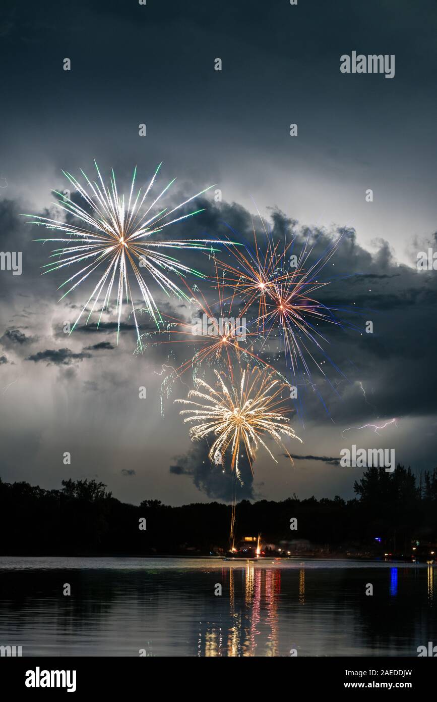 Fireworks explode in front of lightning-illuminated clouds above a lake. Stock Photo