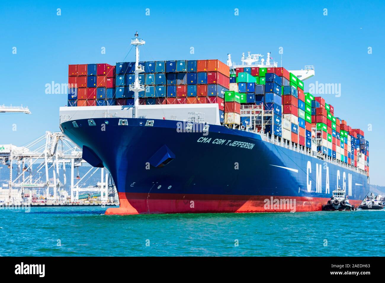 Tugboat assists container ship CMA CGM out of the Port of Oakland under blue sky. A tugboat maneuvers vessel by pushing, pulling or towing containersh Stock Photo