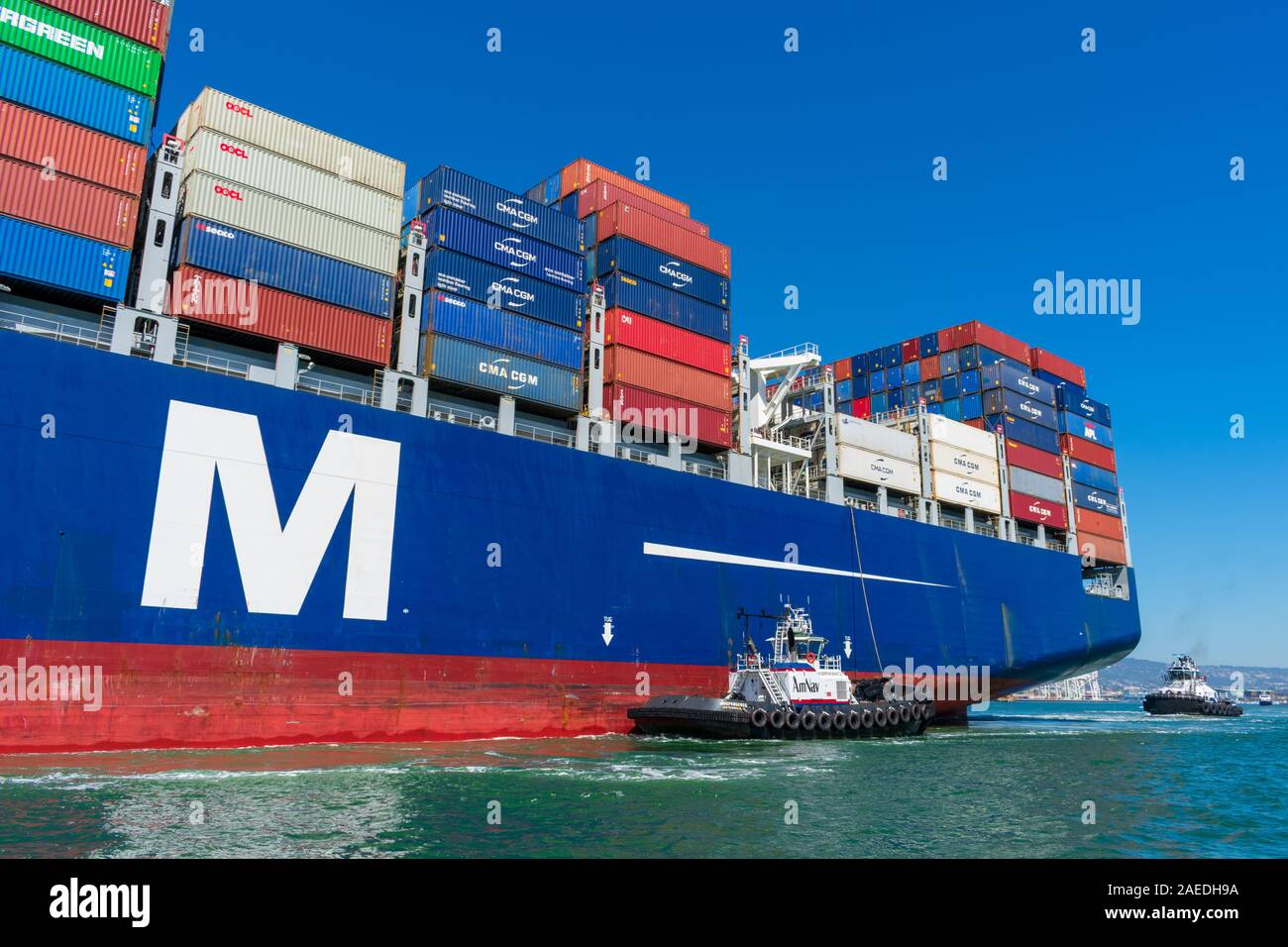 Tugboat assists container ship CMA CGM out of the Port of Oakland under blue sky. A tugboat maneuvers vessel by pushing, pulling or towing containersh Stock Photo
