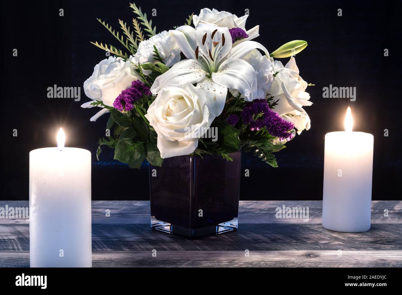 Bouquet of white flowers in a purple vase, white candle on a wooden boards. Vintage home decor dark tones. Condolence card. Stock Photo