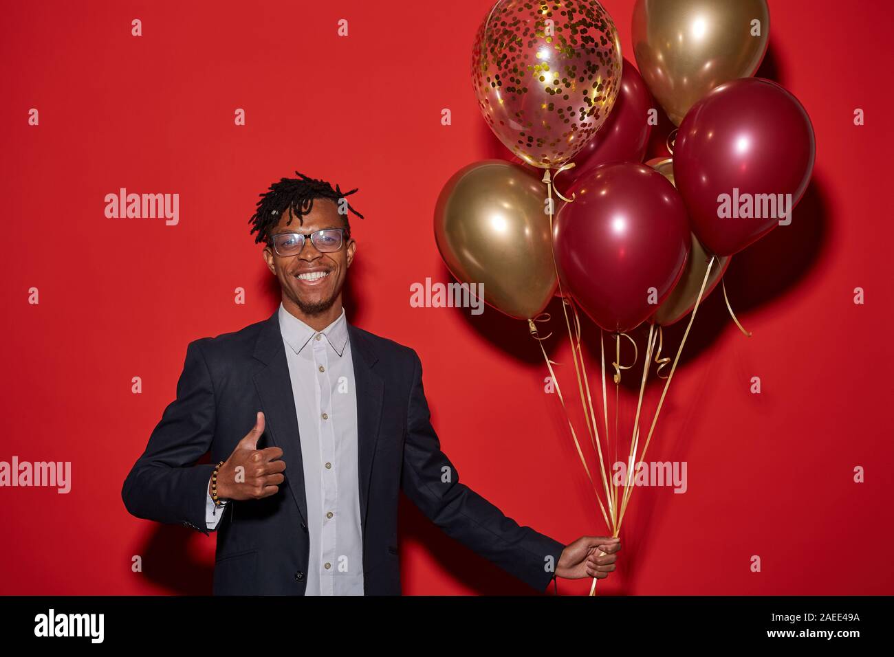 Waist up portrait of smiling African man showing thumbs up and holding bunch of golden balloons while standing against red background at party, copy space Stock Photo
