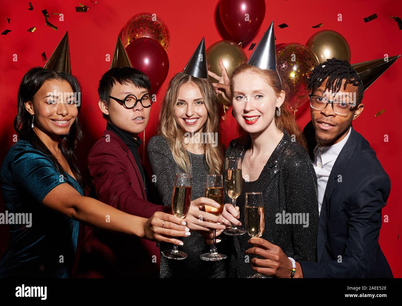 Multi-ethnic group of young people clinking champagne glasses while posing over red background enjoying party, shot with flash Stock Photo