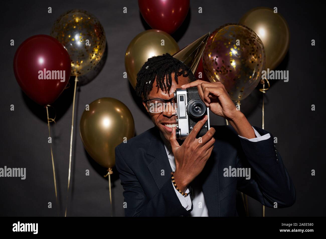 Waist up portrait of contemporary African man holding photo camera while posing against black background with party balloons, shot with flash Stock Photo
