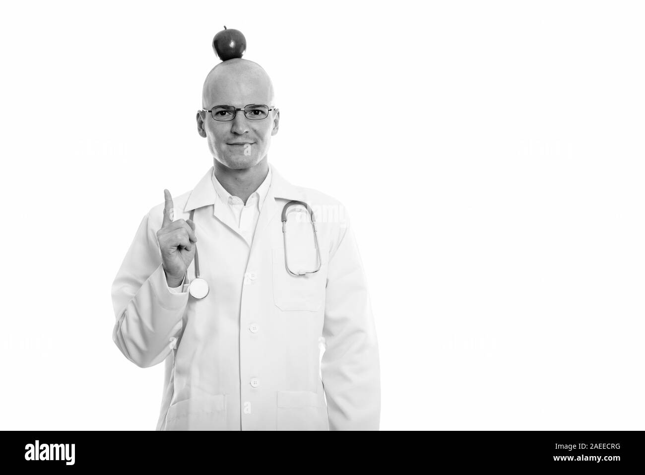 Studio shot of young handsome bald man doctor pointing finger up with red apple on top of his head Stock Photo