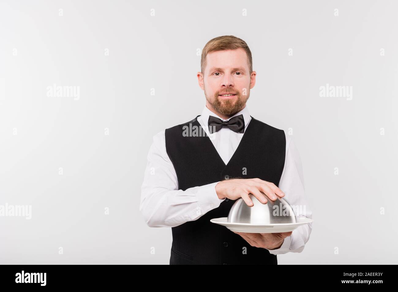 Elegant young waiter in bowtie and black waistcoat holding cloche with food Stock Photo