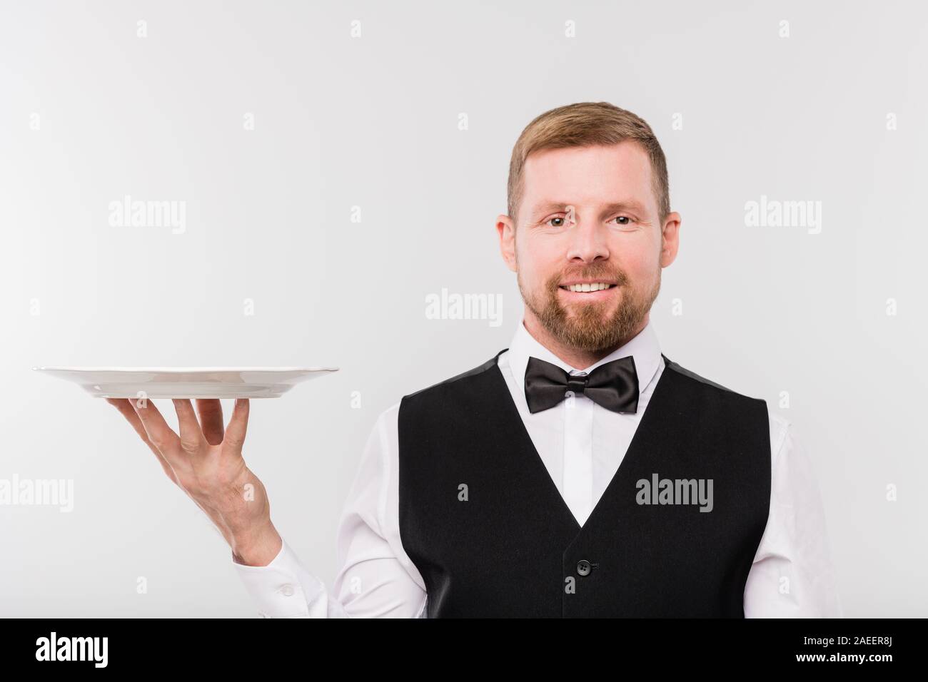 Happy young waiter in bowtie and black waistcoat holding white clean plate Stock Photo