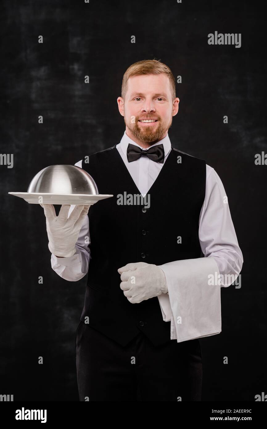 Gloved young waiter holding cloche with food and white towel for one of clients Stock Photo