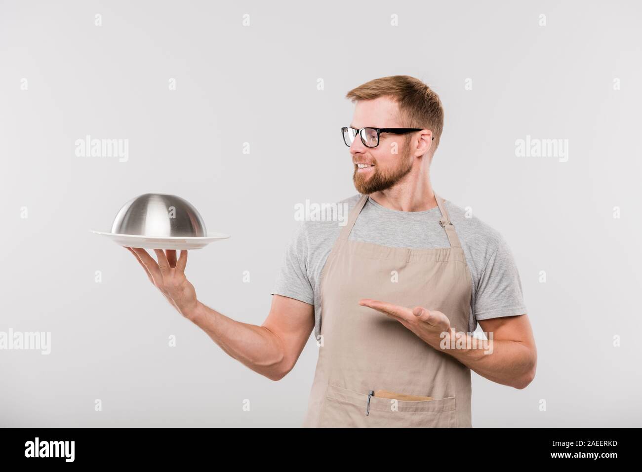 Happy young bearded waiter of cafe in apron and eyeglasses showing cloche Stock Photo
