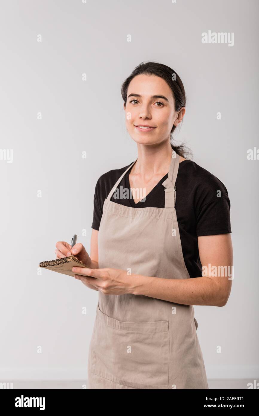 Pretty brunette waitress of cafe in apron making notes in small notepad Stock Photo