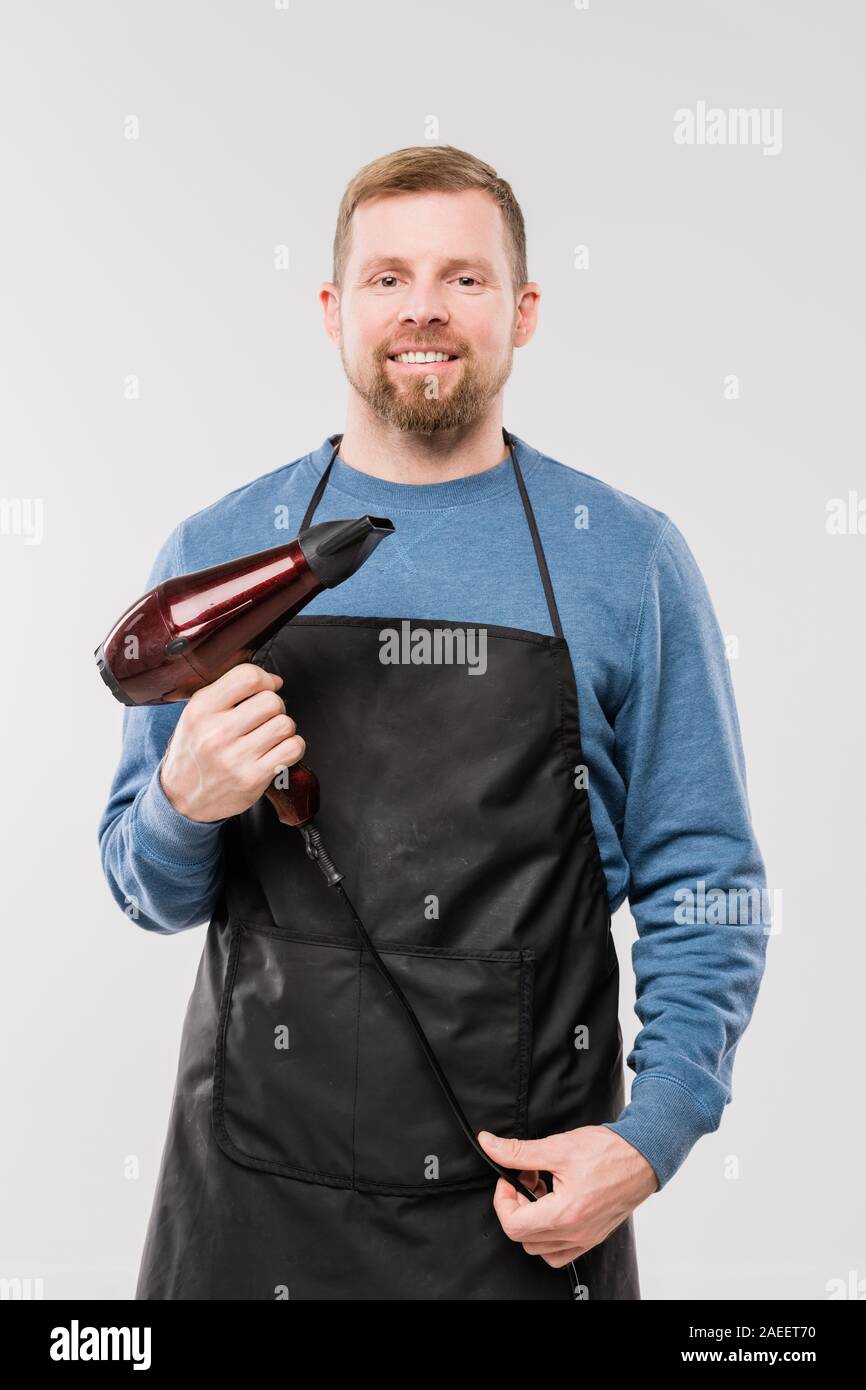 Happy young hairdresser in apron and pullover holding hairdryer while working Stock Photo