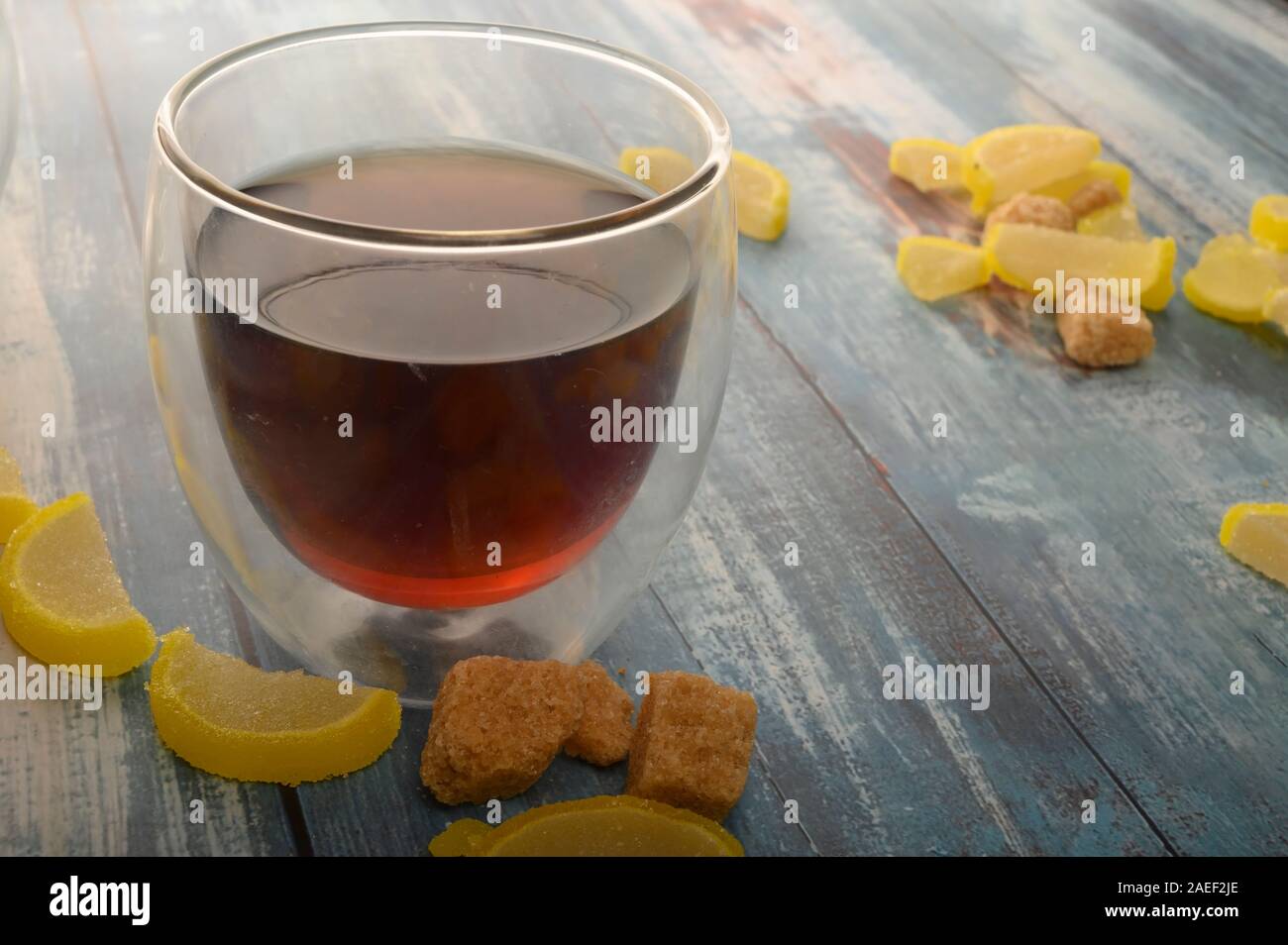 Marmalade lemon slices, pieces of brown sugar and a glass of black tea on a wooden background. Sweet dessert. Close up Stock Photo