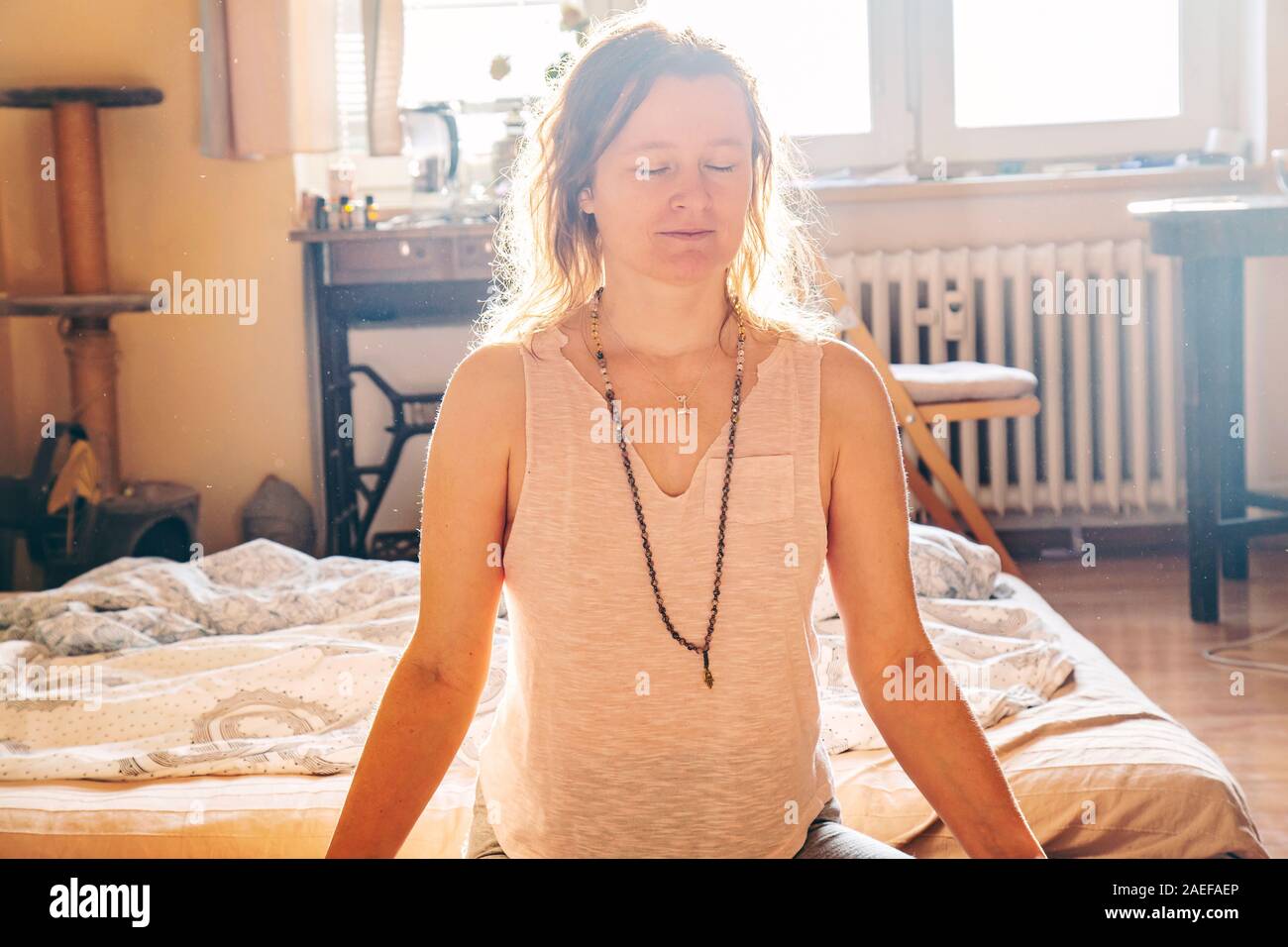 Young yogini woman boho woman with mala necklace meditating in lotus asana in sunny room Stock Photo