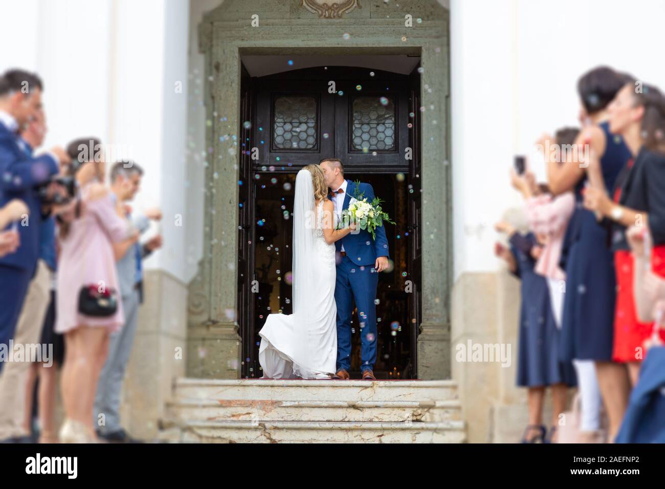 Newlyweds kissing while exiting the church after wedding ceremony, family and friends celebrating their love with the shower of soap bubbles, custom Stock Photo