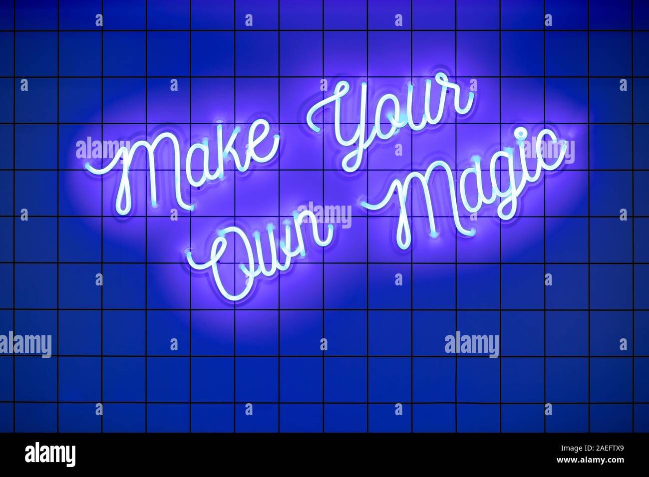 Handwritten neon glowing text Make Your Own Magic on blue tiled wall. Trendy classic color. Luminescent illuminated lamp Stock Photo