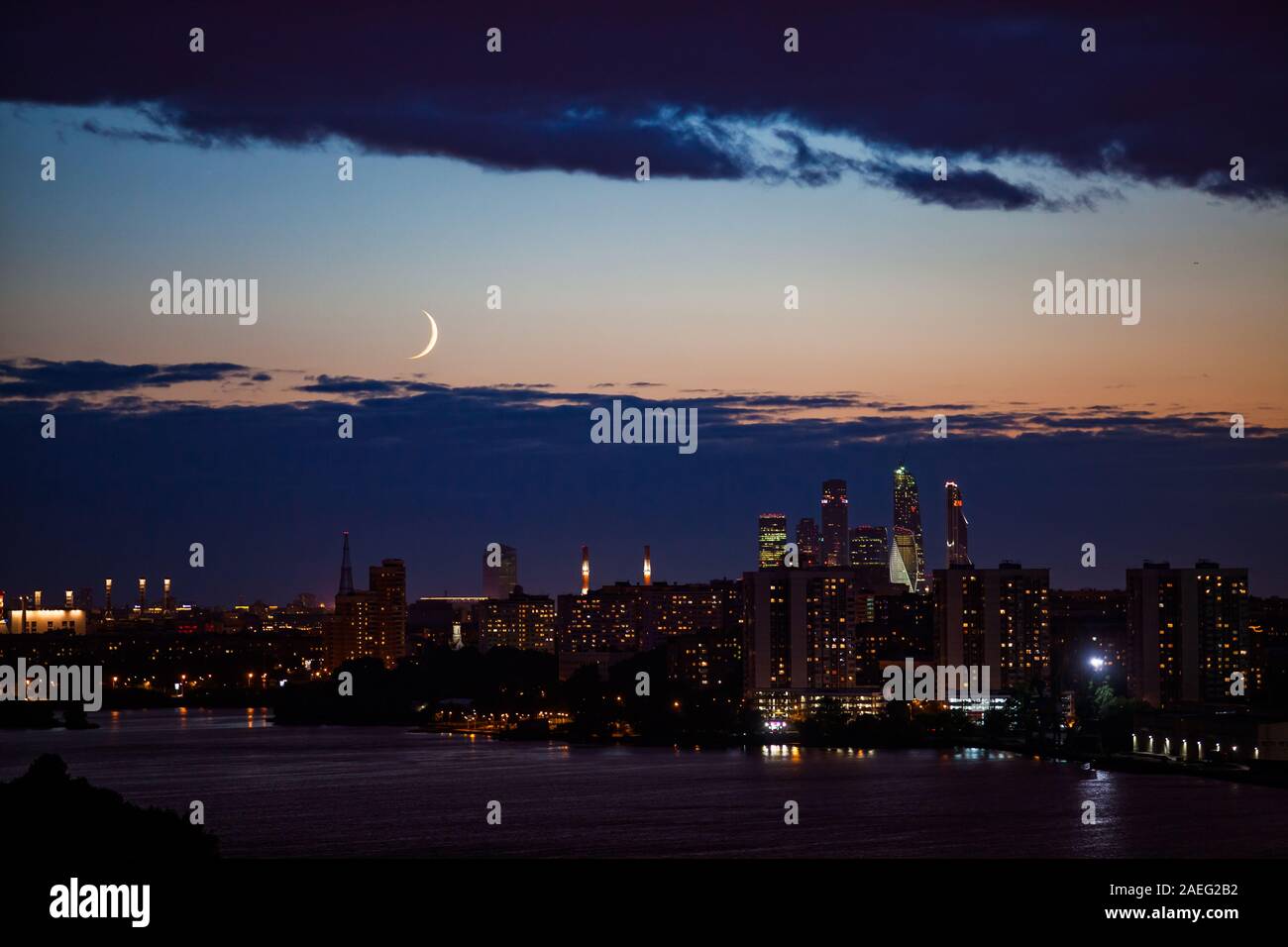 Night or evening city scape with skyscrapers and town silhouette on river in deep blue and yellow sky with clouds and crescent. Stock Photo