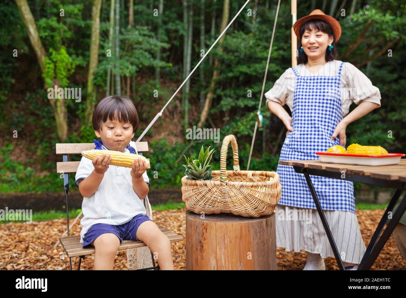 Japanese woman standing outdoors, wearing hat and apron and young boy sitting on chair, eating corn on the cob. Stock Photo
